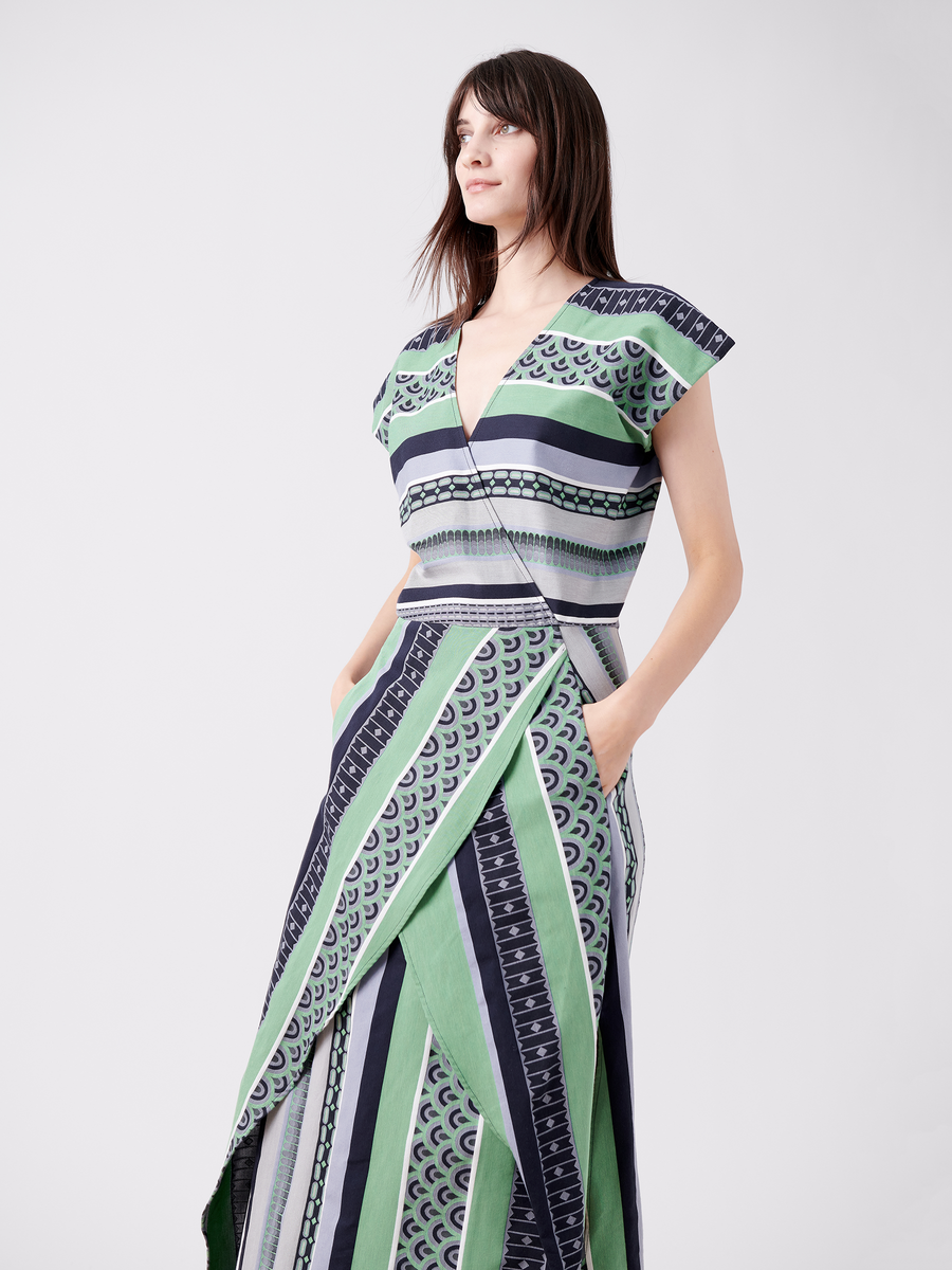 A woman models the Mido Dress by Zero + Maria Cornejo. This long jacquard dress, featuring green, blue, and white geometric stripes, is crafted from organic cotton and includes short sleeves, a wrap-effect V-neckline, and pockets as she poses against a plain white background.