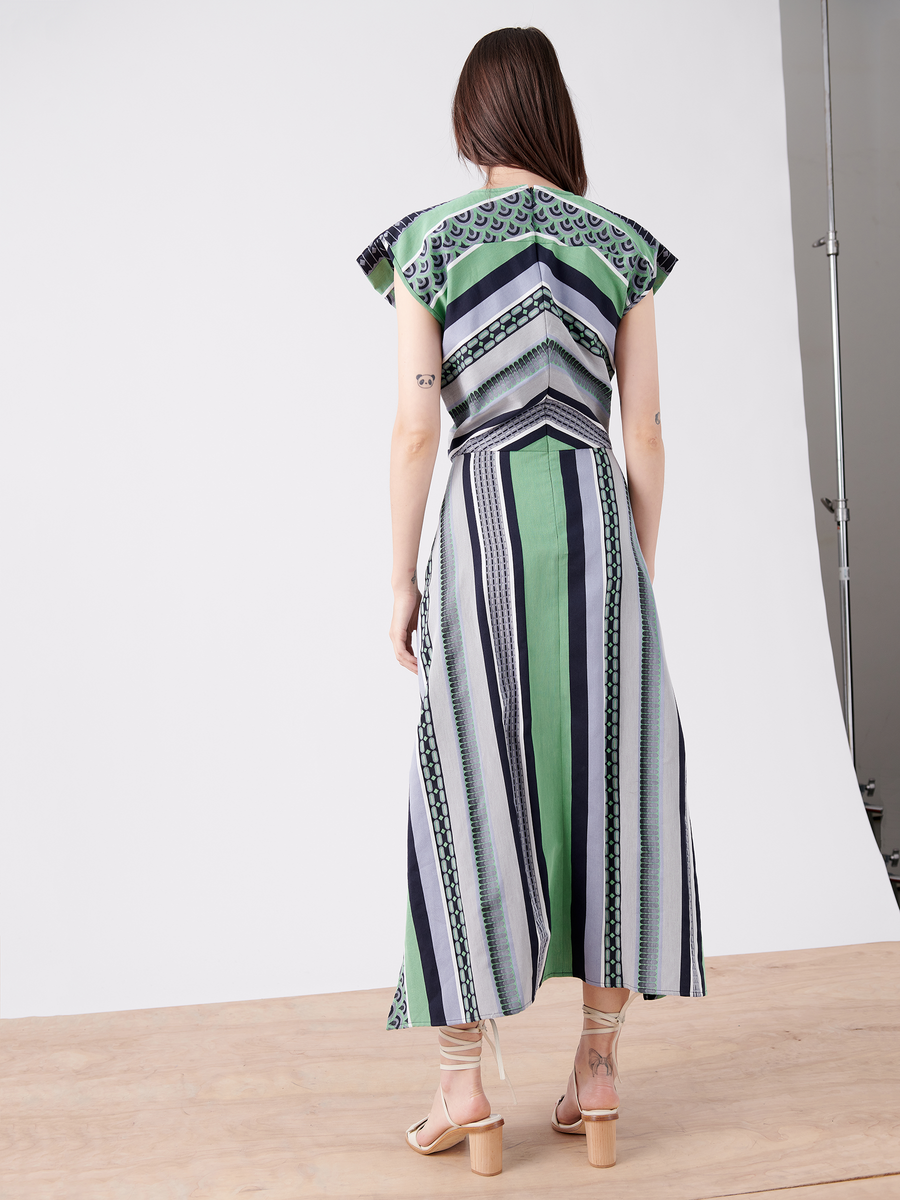 A woman with long brown hair stands on a wooden floor, facing away. She wears the Mido Dress from Zero + Maria Cornejo, a green and blue organic cotton wrap-effect v-neck dress, and beige block-heeled sandals in a white studio setting.