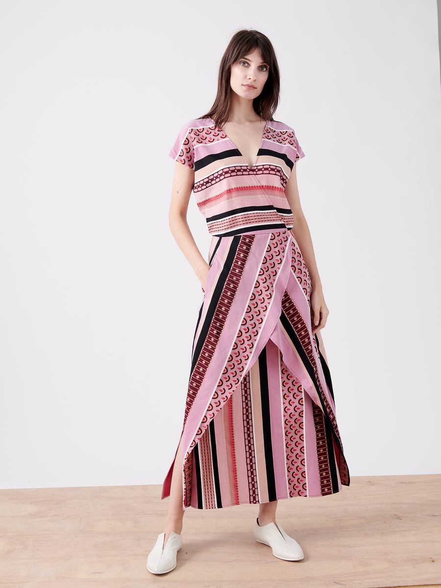 Wearing the Mido Dress by Zero + Maria Cornejo, the person showcases a vibrant geometric-patterned dress made from organic cotton. The striped jacquard fabric features pink, red, and black hues with a wrap-effect V-neckline. They're on a wooden floor with white slip-on shoes against a plain white background.