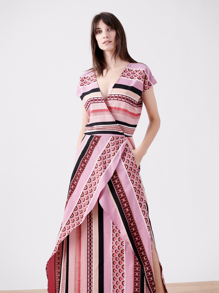 A woman models the Mido Dress by Zero + Maria Cornejo, featuring pink, red, and black stripes with geometric designs. The striped jacquard dress includes short sleeves, a wrap-effect V-neck bodice, and side pockets as she poses against a plain background.