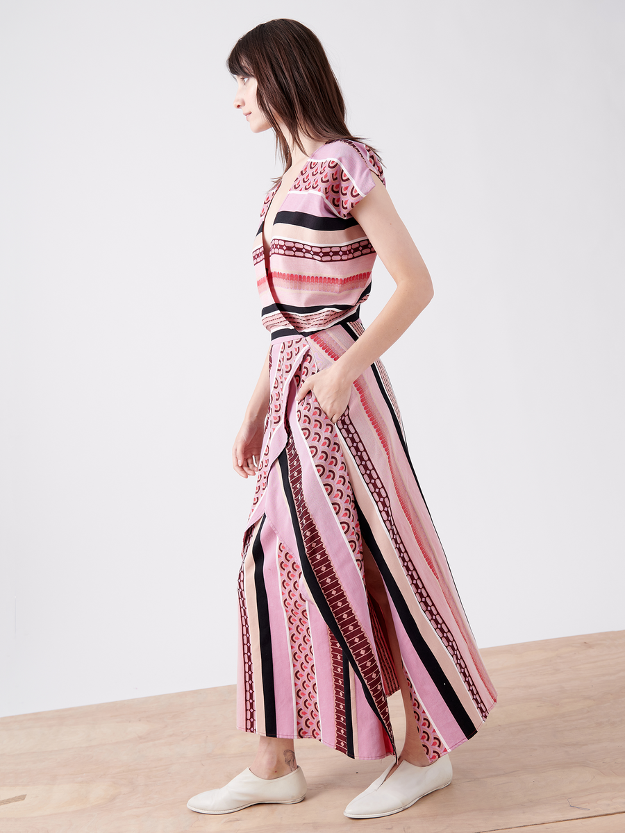 A woman with long brown hair wears the Mido Dress from Zero + Maria Cornejo, a pink striped jacquard design with an elegant wrap-effect V-neck. She stands sideways, her left hand in the pocket of this organic cotton dress, paired with white slip-on shoes against a plain background.