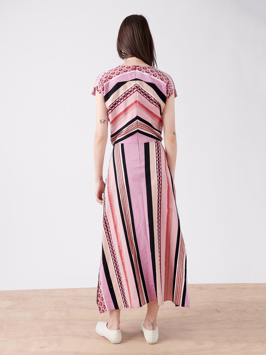 A person with long hair wears the Mido Dress - Final Sale by Zero + Maria Cornejo, a pink and black striped jacquard dress with geometric patterns. The wrap-effect V-neck adds elegance as they stand on a wooden floor against a white background, paired with white sneakers.