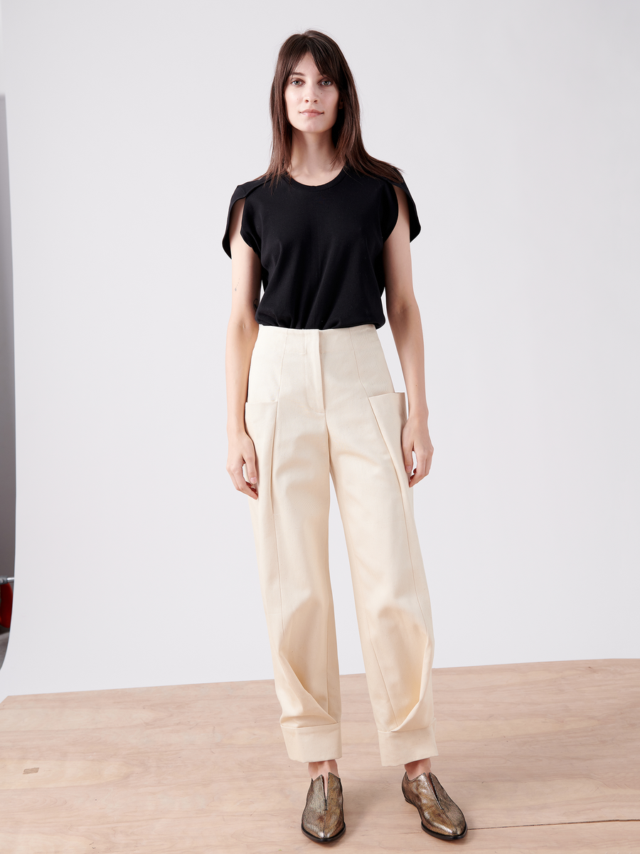 A person with long hair is on a wooden platform wearing a black T-shirt, light beige Zero + Maria Cornejo Jodhpur trousers (Cradle 2 Cradle Certified), and metallic bronze slip-on shoes against a plain white backdrop.