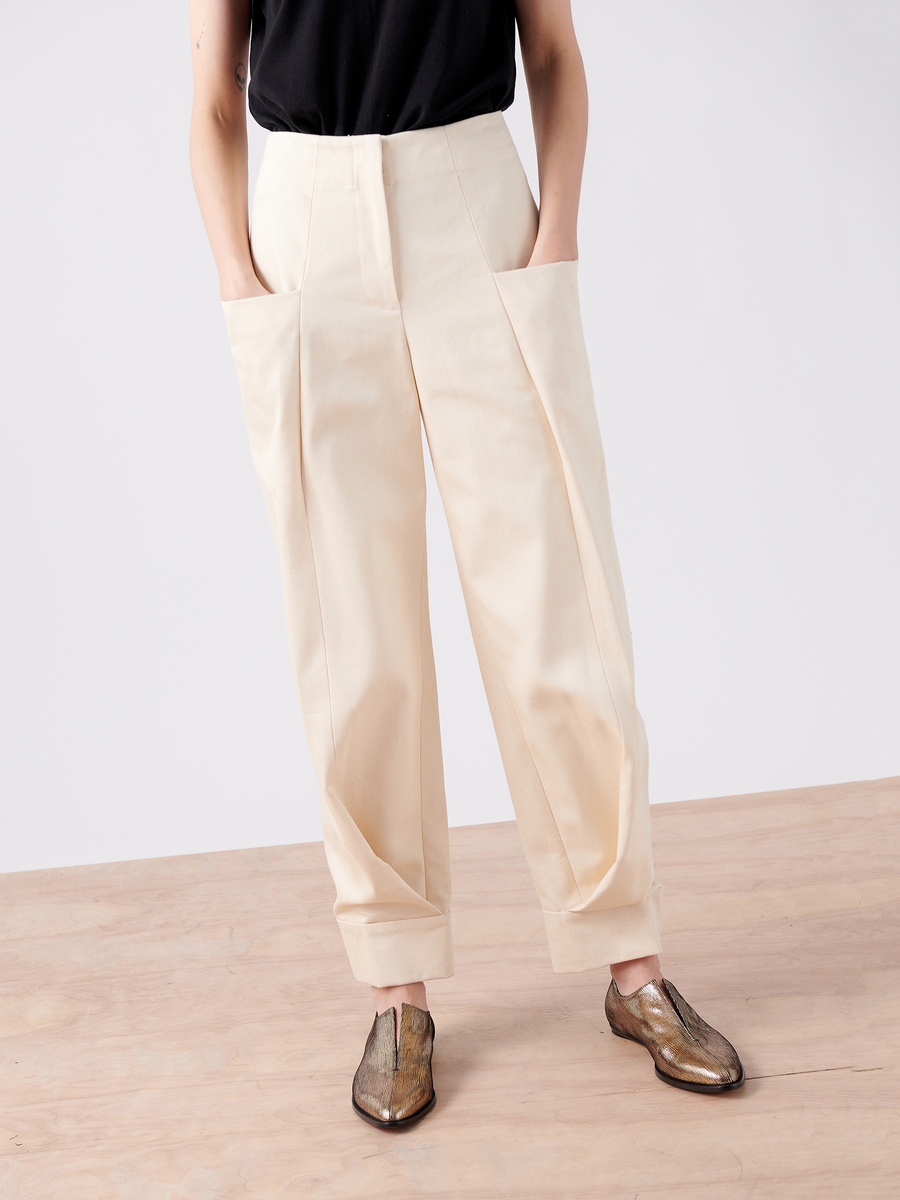 A person wears cream-colored Akeo Jodhpur pants from Zero + Maria Cornejo, featuring wide legs, large front pockets, and 100% organic cotton. Styled with a black top and metallic brown shoes against a minimalist backdrop, the Cradle 2 Cradle Certified pants blend style with sustainability.