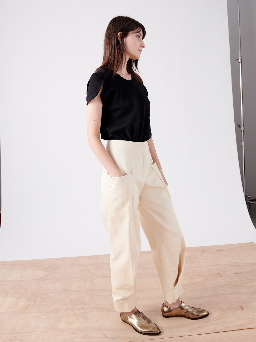 A person with long dark hair stands on a wooden floor against a plain backdrop, wearing a black shirt, Cradle 2 Cradle Certified beige pants, and metallic gold Akeo Jodhpur shoes by Zero + Maria Cornejo, with hands in pockets and looking to the side.