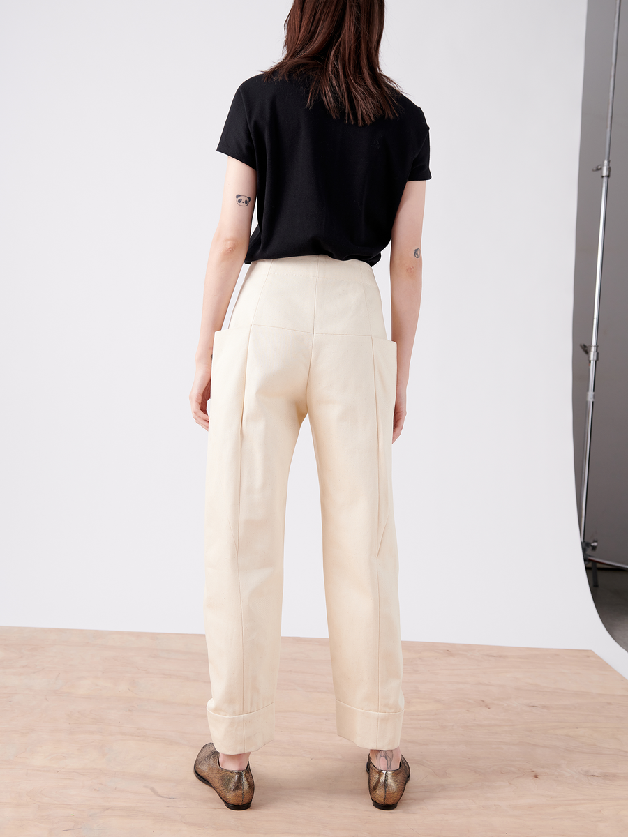 A person faces away from the camera, dressed in a black T-shirt, Cradle 2 Cradle Certified high-waisted cream pants, and Akeo Jodhpur - Final Sale by Zero + Maria Cornejo. The shot is set in a studio featuring a wooden floor and a white backdrop.