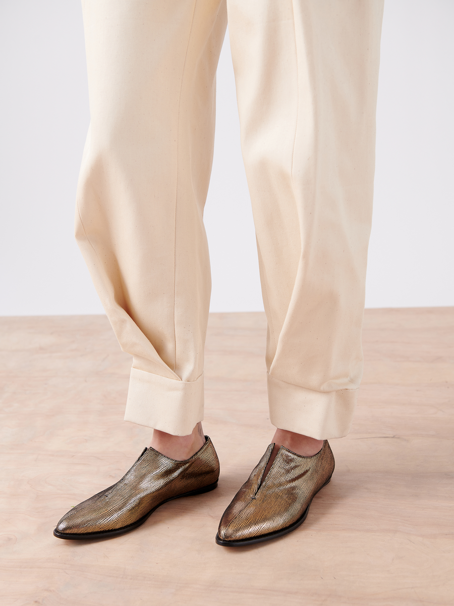 A person in cream cuffed pants stands on a wooden floor, showcasing metallic brown Akeo Jodhpur slip-ons with a slight shimmer by Zero + Maria Cornejo.