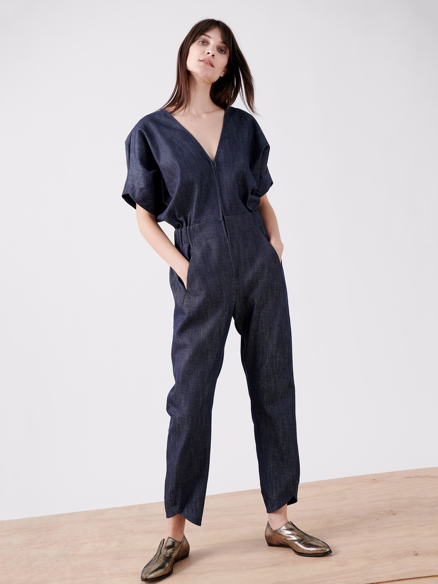 On a wooden floor, someone wears the Aissa V-Neck Jumpsuit by Zero + Maria Cornejo in dark blue organic denim, featuring an elastic waist and short sleeves. They pair it with metallic gold shoes and stand with hands in pockets against a plain white background, looking ahead.