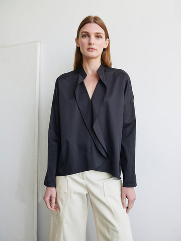 A person with long brown hair stands against a plain white background, wearing a Zero + Maria Cornejo Long-Sleeved Fin Gaban Shirt and light-colored pants. Their hands are relaxed by their sides, and they have a calm expression on their face.