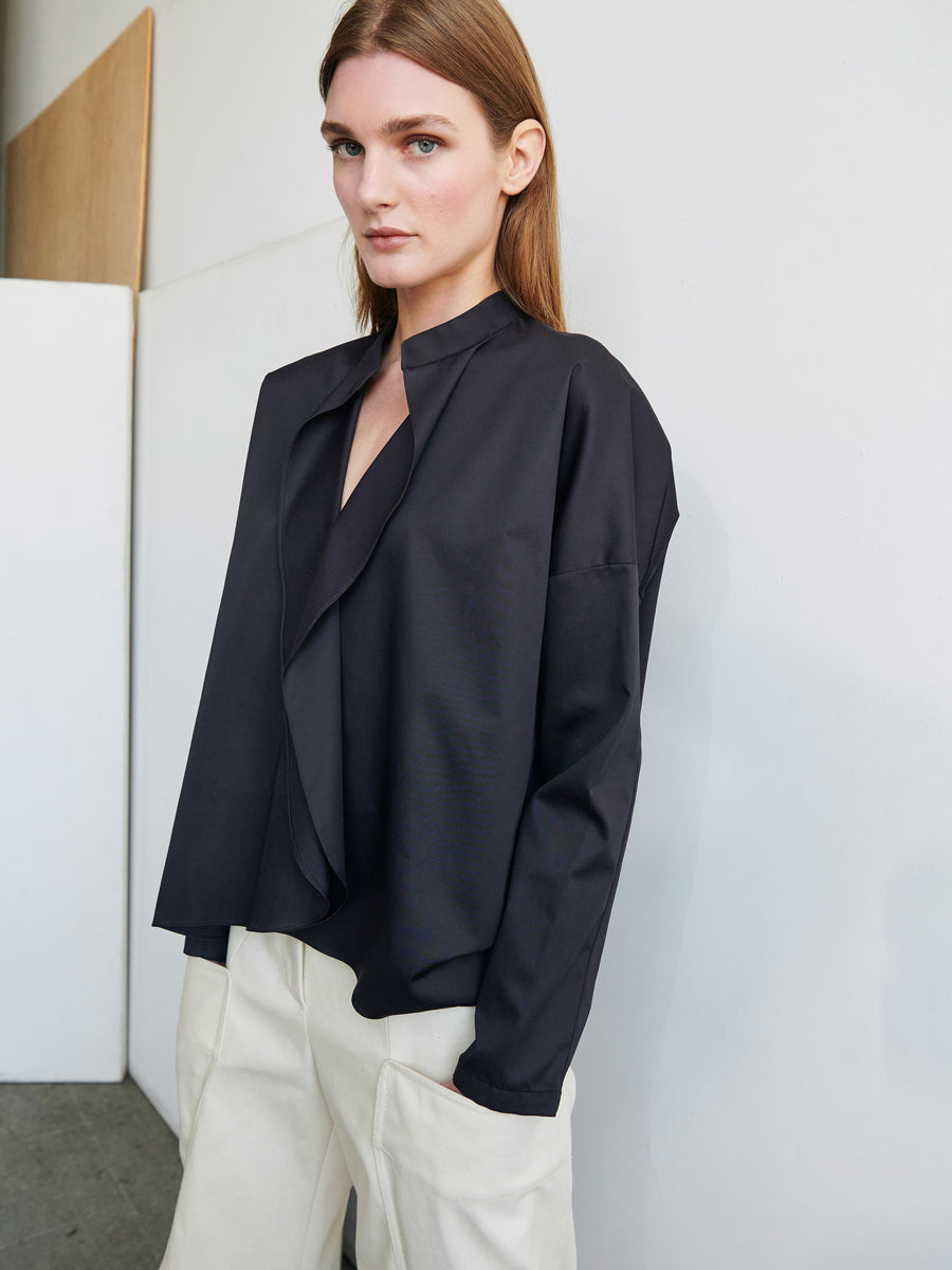 A person stands indoors against a plain wall, wearing a black Long-Sleeved Fin Gaban Shirt by Zero + Maria Cornejo and loose-fitting beige pants. They have shoulder-length straight brown hair and a neutral expression. The setting appears minimalist and modern, reminiscent of the simplicity of a cotton broadcloth shirt.