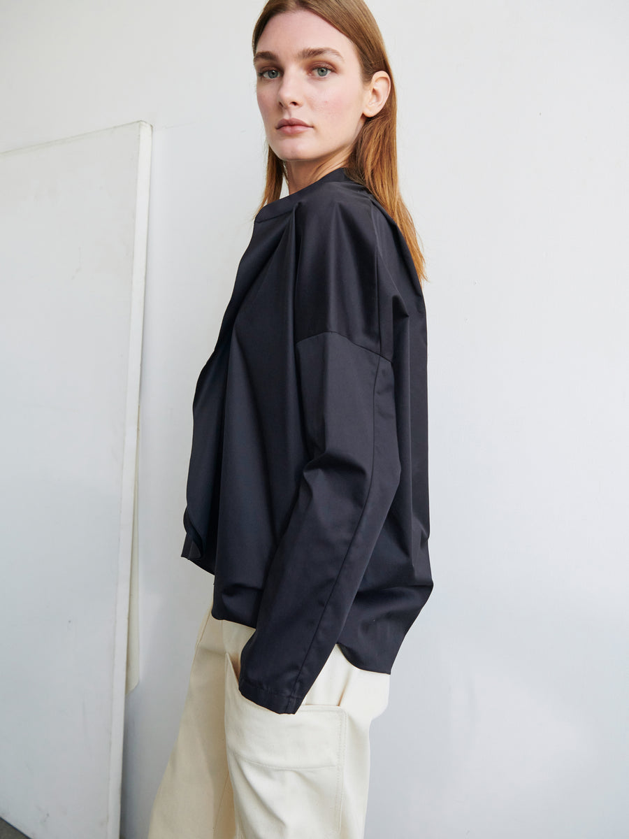 A person with long, straight hair is wearing a loose-fitting black jacket over a light-colored Long-Sleeved Fin Gaban Shirt by Zero + Maria Cornejo and matching pants. They are standing in front of a white wall, looking towards the camera with a neutral expression.