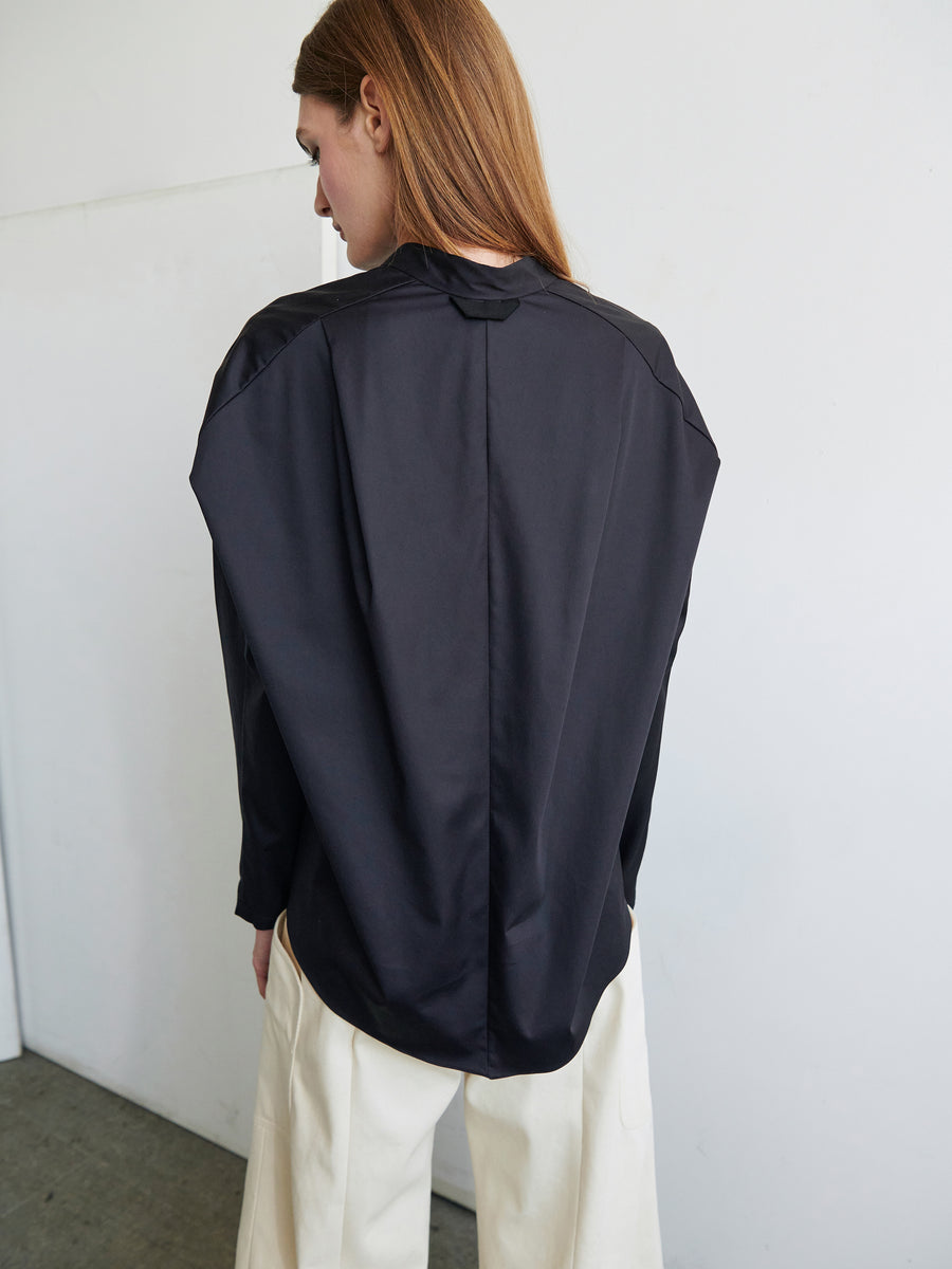 A person with long, light brown hair stands facing a white wall, wearing a Long-Sleeved Fin Gaban Shirt by Zero + Maria Cornejo and white pants. The focus is on their back, highlighting the sleek and minimalist design of the cotton broadcloth shirt.