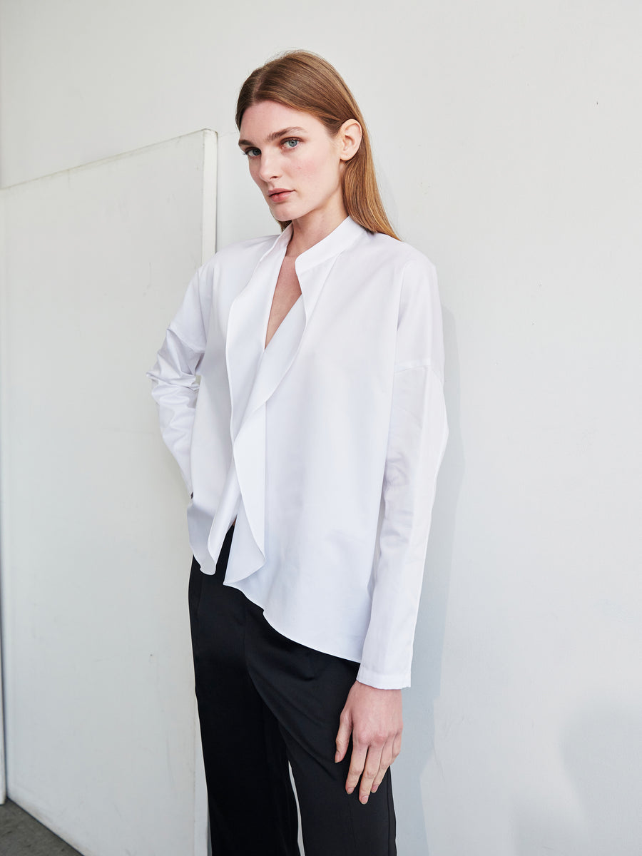 A person with long, light brown hair stands against a blank wall. They are wearing a loose-fitting, white Long-Sleeved Fin Gaban Shirt made of cotton broadcloth from Zero + Maria Cornejo and dark pants. Their left hand rests on their hip, and they have a calm, neutral expression.