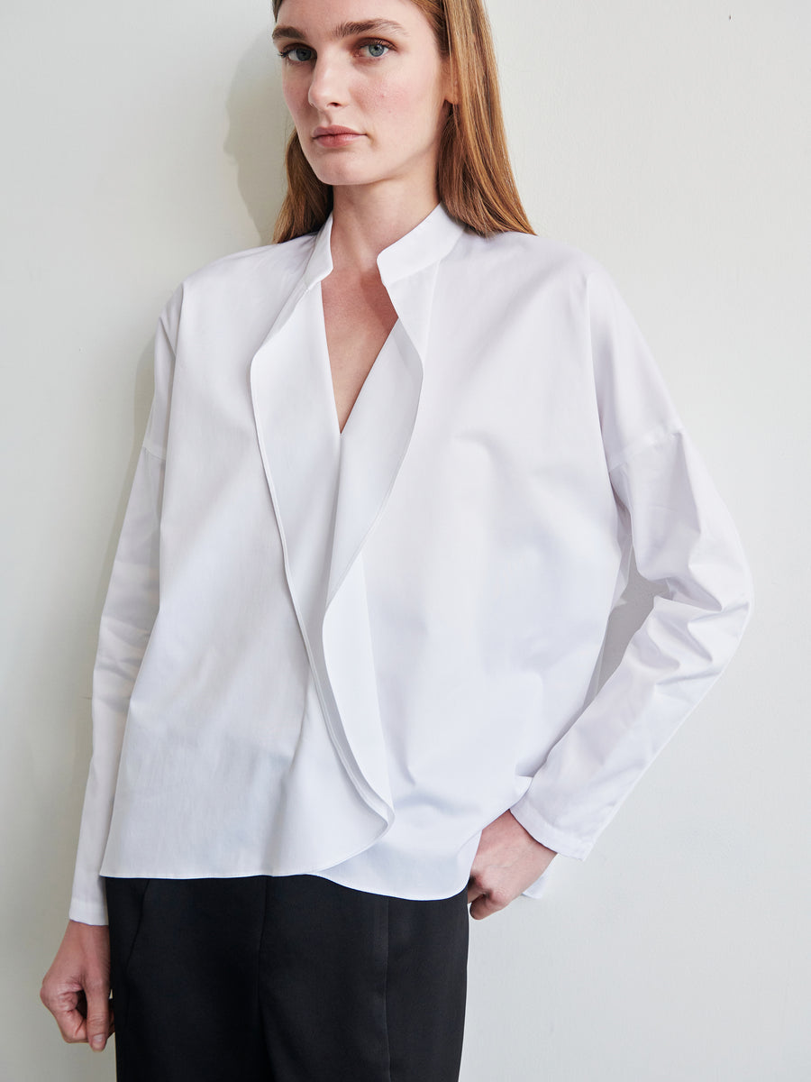 A person stands against a plain background, wearing a Long-Sleeved Fin Gaban Shirt by Zero + Maria Cornejo paired with black trousers. The shirt features an open collar and is made of white cotton broadcloth. They have straight, shoulder-length hair and a neutral expression, with one hand resting by their side and the other in their pocket.