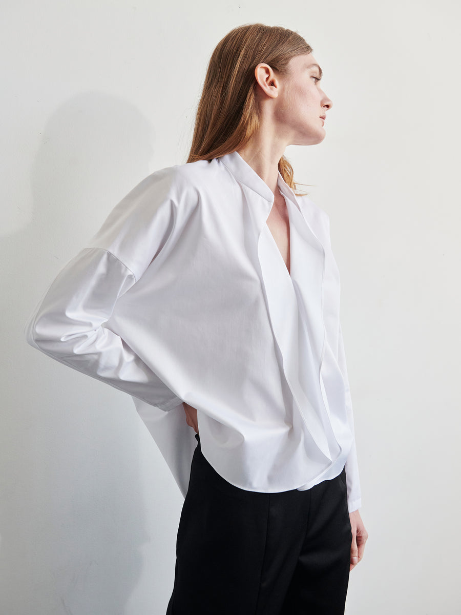 A person with long hair is wearing a Long-Sleeved Fin Gaban Shirt by Zero + Maria Cornejo, featuring a draped design with a deep V-neck. They are paired with loose-fitting black pants and are standing against a plain white background, looking to the side with one hand in their pocket. A stand collar adds an element of sophistication to their look.