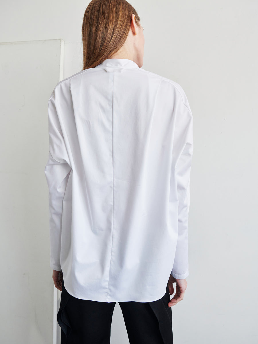 A person with long hair is seen from behind, wearing a white Long-Sleeved Fin Gaban Shirt by Zero + Maria Cornejo, characterized by a draped ruffle neckline. The background is plain and minimalist, featuring a light-colored wall.