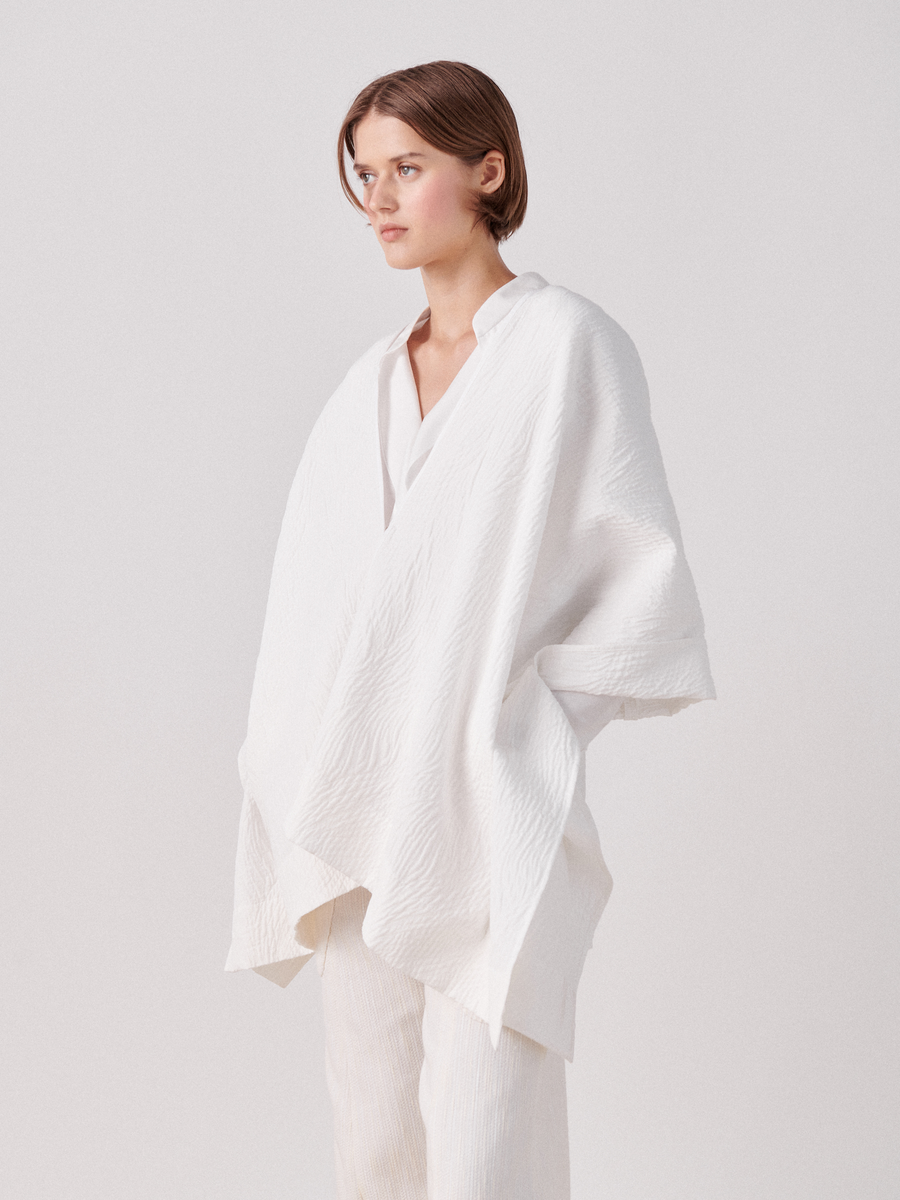 A person with short brown hair stands against a plain background, wearing a loose-fitting white outfit that includes a flowing top and matching pants made of organic cotton. They complement the look with an Eve Square Shrug by Zero + Maria Cornejo and are looking to the right with a neutral expression.
