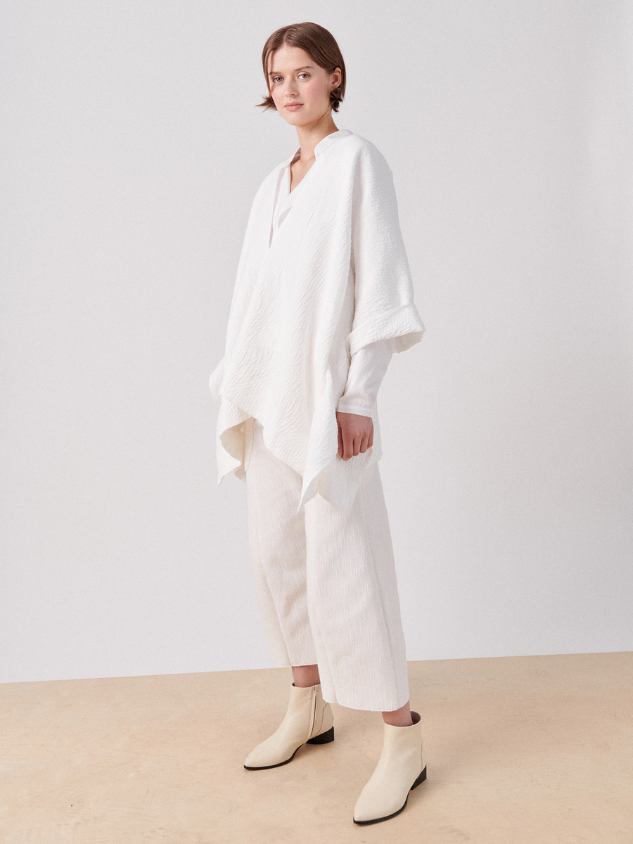 A person with short hair stands in a minimalistic setting, wearing the Eve Square Shrug from Zero + Maria Cornejo, paired with white ankle-length pants. They are also wearing light-colored, pointed-toe ankle boots and looking towards the camera.