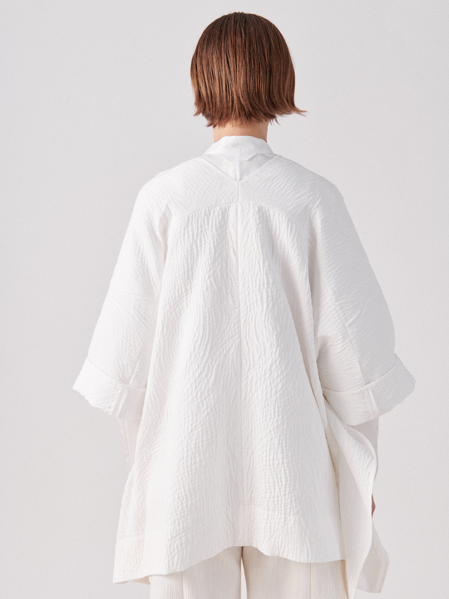 A person with short brown hair stands with their back to the camera, wearing an oversized, textured white garment. This Eve Square Shrug by Zero + Maria Cornejo features wide sleeves and appears to be made of lightweight fabric. The background is plain and white.