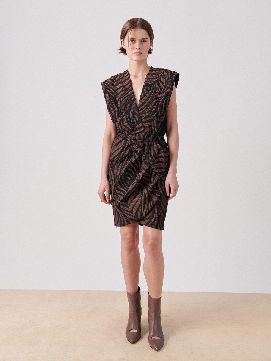 A person stands against a plain white background, wearing the Mackie Dress from Zero + Maria Cornejo, a black and brown patterned wrap v-neck sleeveless dress with a deep V-neck and asymmetric gathering at the waist. They are also wearing brown ankle boots with a pointed toe.