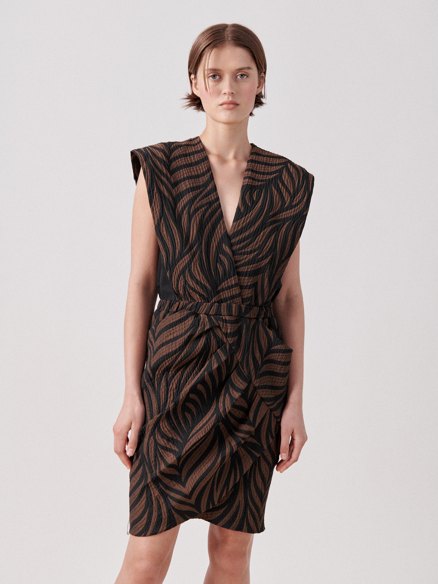 A person with short brown hair stands facing the camera, wearing a sleeveless wrap v-neck Mackie Dress by Zero + Maria Cornejo with a black and brown wavy pattern. The quilted above-knee dress features a belted waist and ruffled detailing at the front. The background is plain and light-colored.