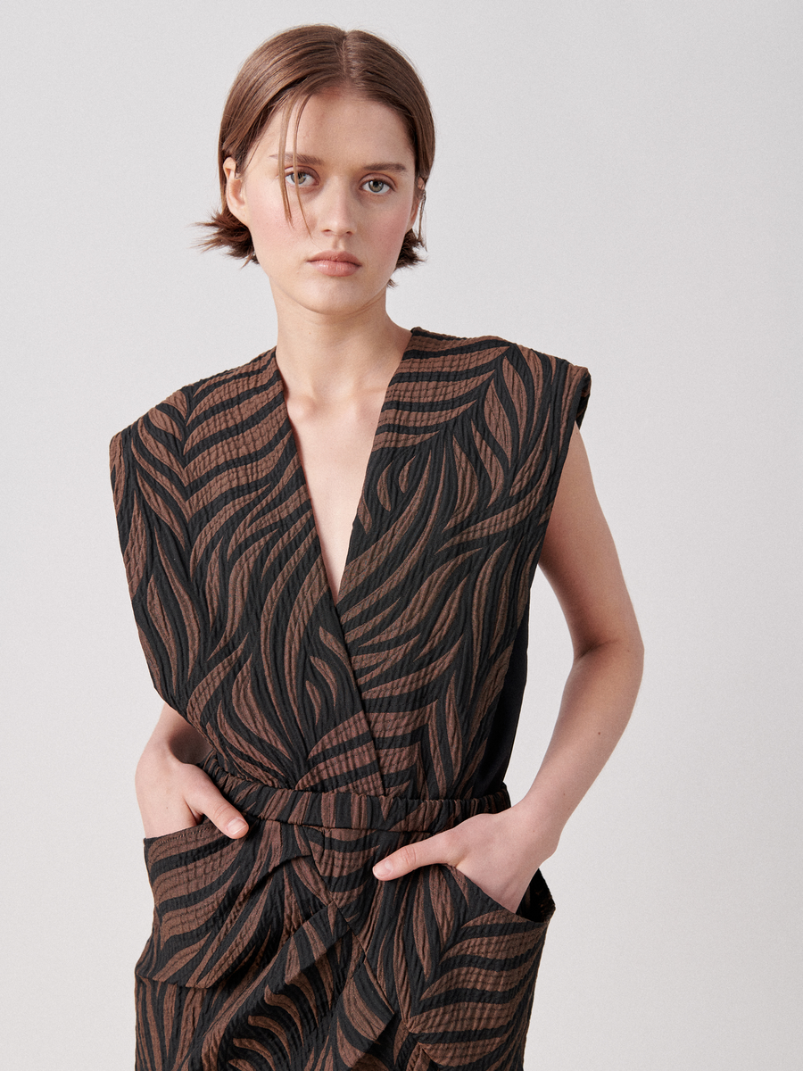 A person with short hair is wearing a sleeveless, wrap V-neck Mackie Dress by Zero + Maria Cornejo with a bold, black and brown leaf pattern. Their hands are in the dress pockets, and they are looking directly at the camera against a plain background.