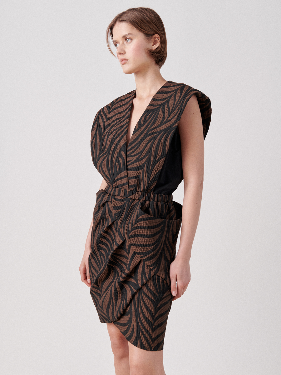 A person with short brown hair is wearing the Mackie Dress by Zero + Maria Cornejo, a sleeveless, structured dress featuring a brown and black leaf pattern in organic cotton. The above-knee dress has a wrap V-neck and an exaggerated shoulder design. The individual is looking to the side against a plain white background.