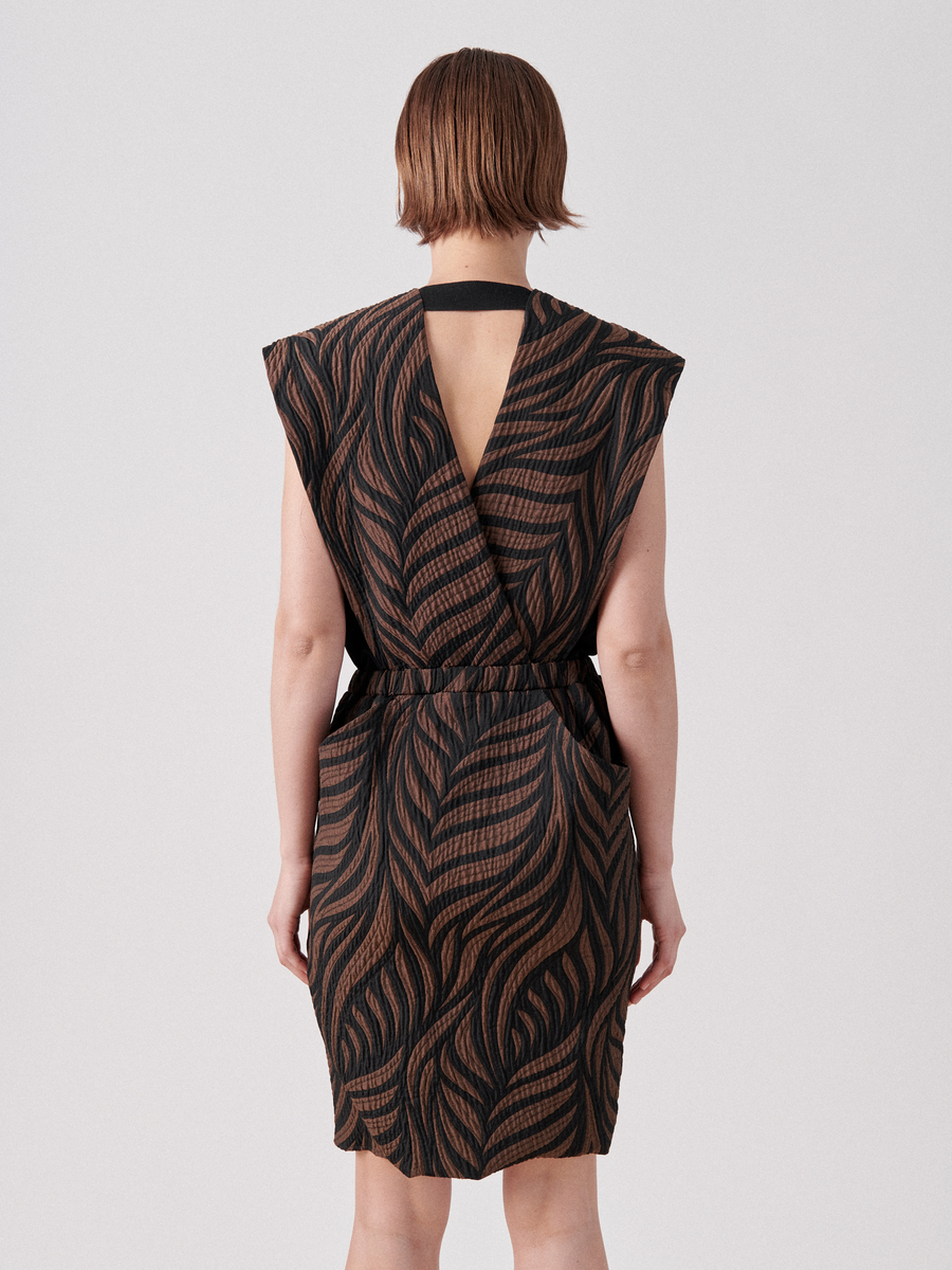 A person stands with their back to the camera, wearing the Mackie Dress by Zero + Maria Cornejo. The quilted above-knee dress features a sleeveless design, organic cotton fabric with a black and brown leaf pattern, a V-shaped back cutout, and a black strap at the neck. The person's hair is short and straight.
