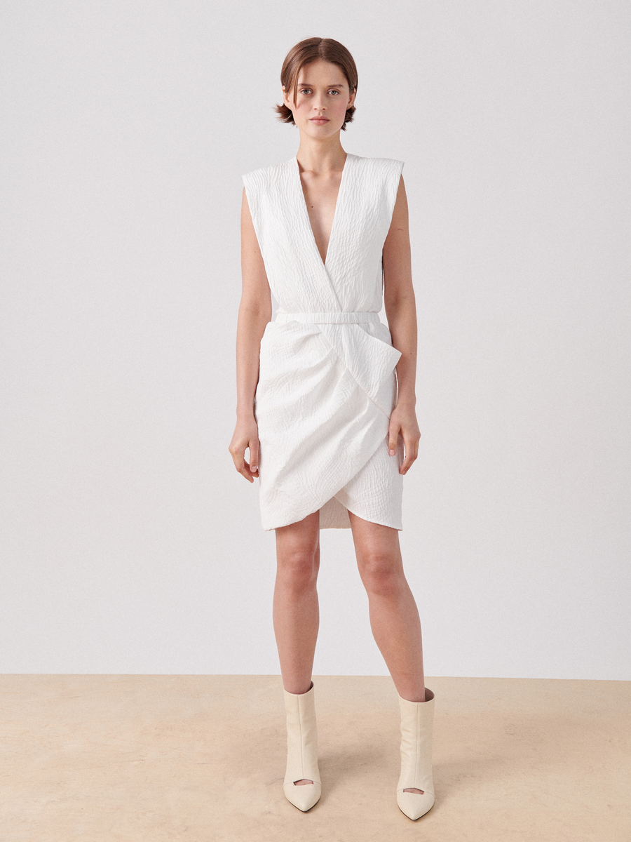 A person stands against a plain white background wearing the Mackie Dress by Zero + Maria Cornejo. The dress is a sleeveless, white textured quilted above-knee dress with a wrap v-neck and structured shoulders. They have short, brown hair and are wearing off-white ankle boots.