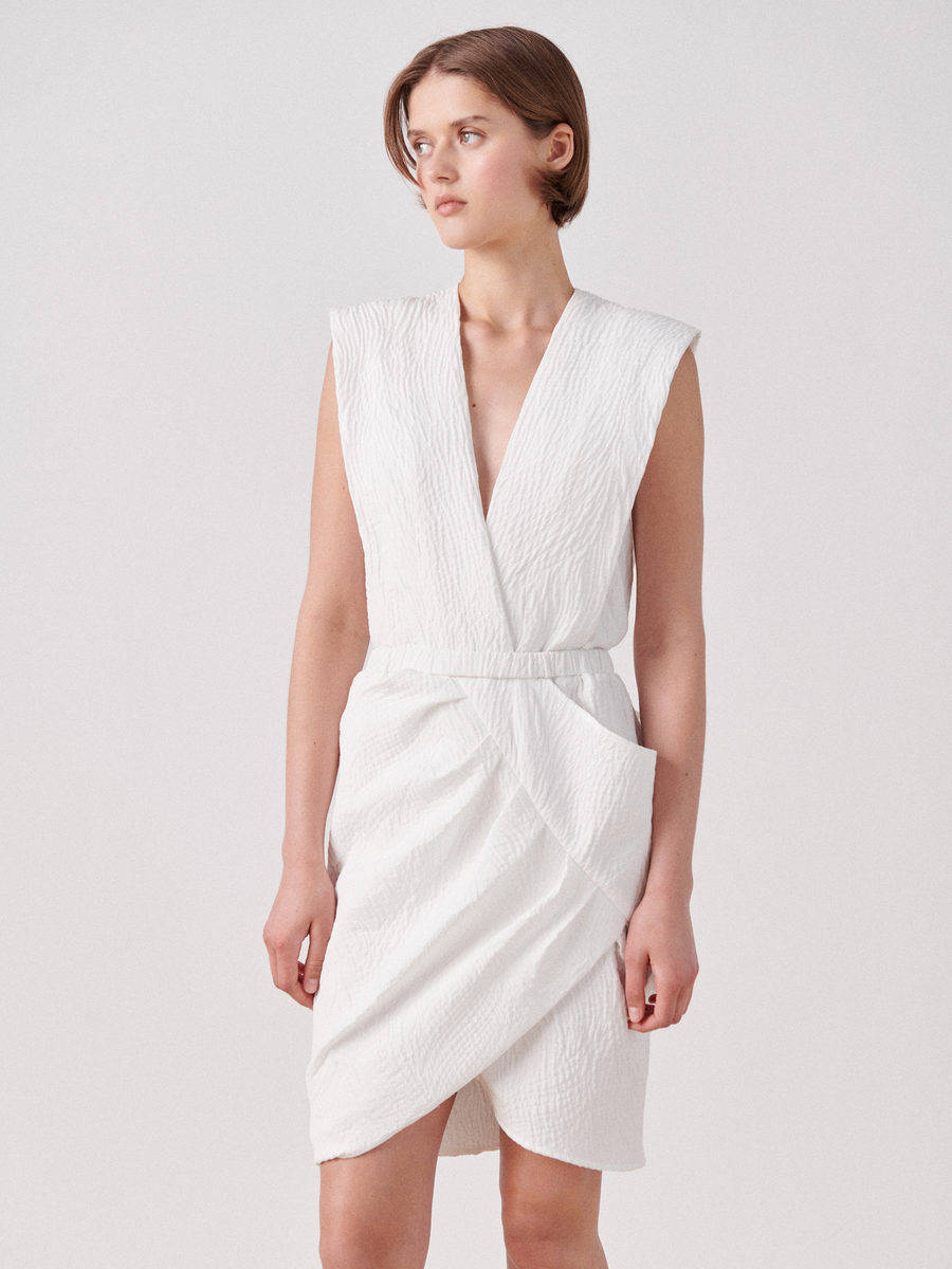 A person with short hair is wearing a Mackie Dress, a sleeveless, white textured dress made from organic cotton by Zero + Maria Cornejo. The wrap V-neck design complements its elegant style, and they stand against a plain background, looking to the side with a neutral expression.