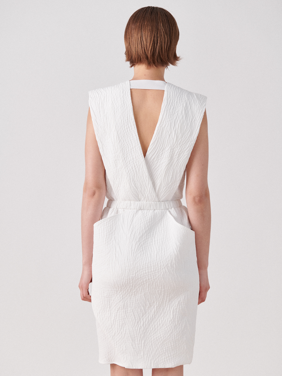 A person with short brown hair is standing with their back to the camera, wearing a sleeveless, white quilted above-knee Mackie Dress from Zero + Maria Cornejo. The dress features a V-shaped cutout at the back and a fitted waistband. The background is plain white.