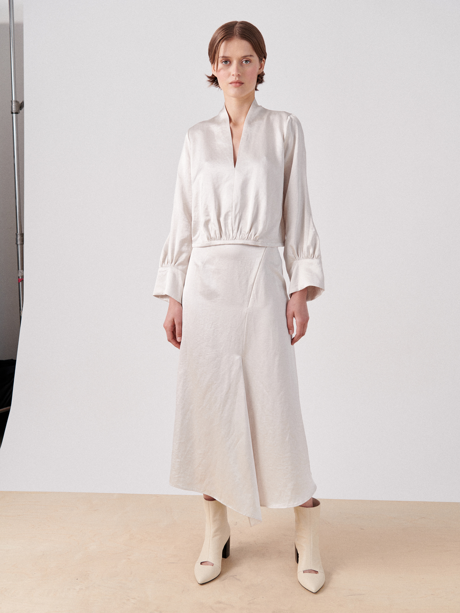 A person with short hair is standing against a plain backdrop. They are wearing a cream-colored, long-sleeved blouse with a plunging neckline and a matching knee-length wrap skirt, the Silent Skirt by Zero + Maria Cornejo, featuring asymmetrical seam details. They also have on white ankle boots. The setting appears to be a studio.