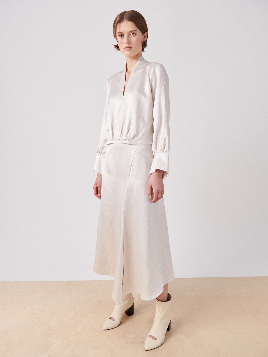 A person with short hair is wearing a light-colored, long-sleeve, midi-length dress with a V-neck and a cinched waist, paired with light-colored ankle boots. They are also wearing the Silent Skirt by Zero + Maria Cornejo. The outfit features an asymmetrical seam detail and an angled hemline. They are standing on a light-colored floor against a plain white background.