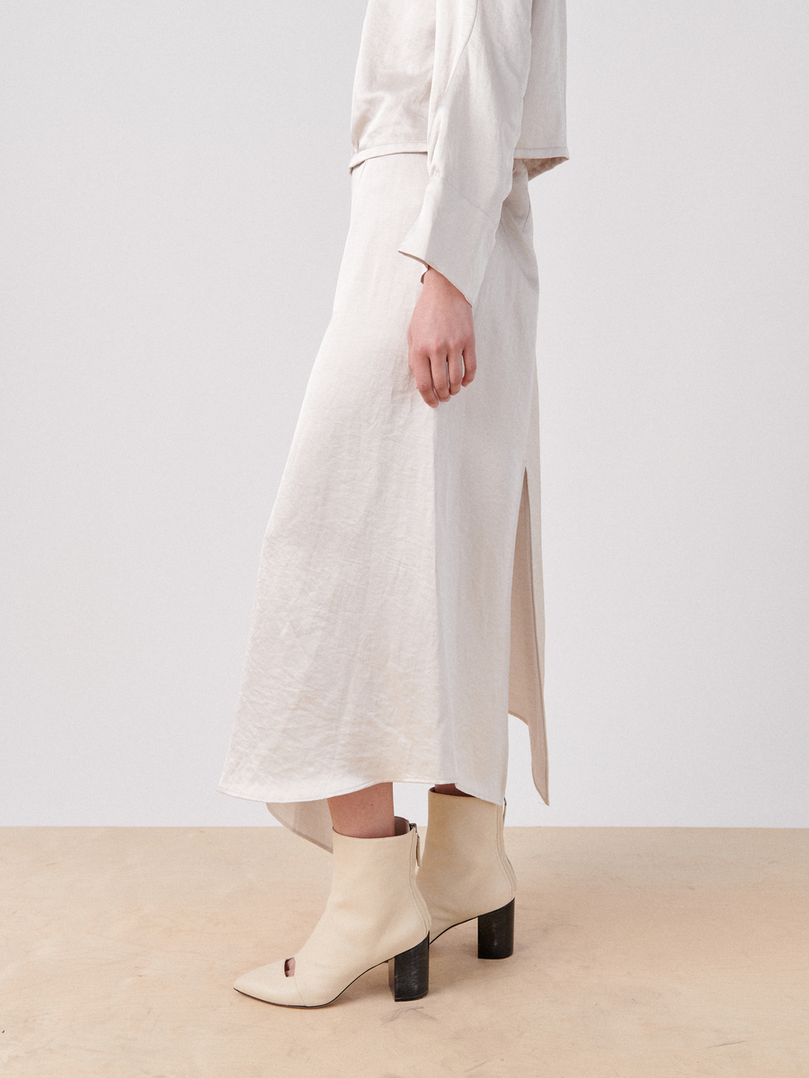 An individual is standing sideways on a light wood floor, wearing an off-white Silent Skirt by Zero + Maria Cornejo with an asymmetrical seam detail that reaches mid-calf and features a side slit. They are also sporting off-white ankle boots with block heels. The background is plain and light-colored.