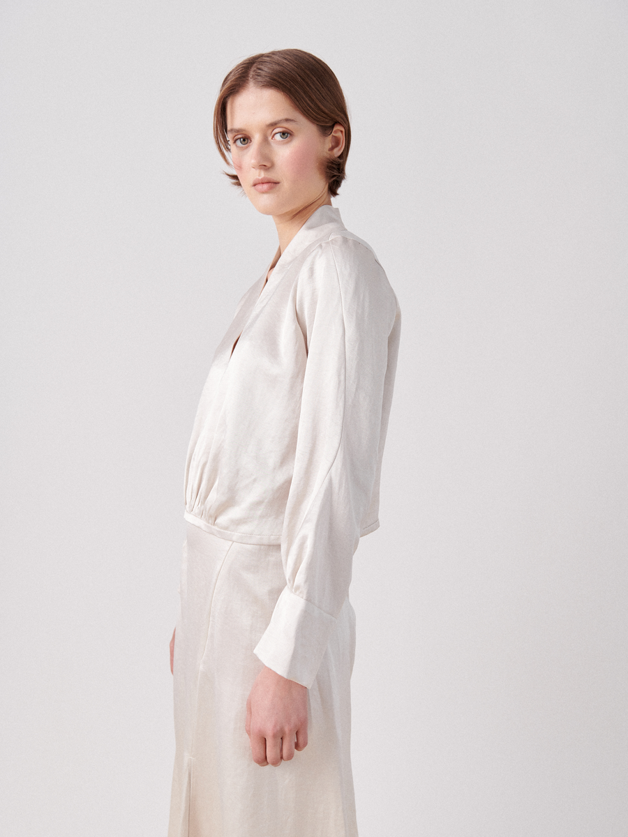 A person with short brown hair stands facing sideways and slightly looking toward the camera. They are wearing a light-colored, Zero + Maria Cornejo Simple Sacha Top with a v-neckline and matching skirt featuring full long sleeves. The background is plain and white.