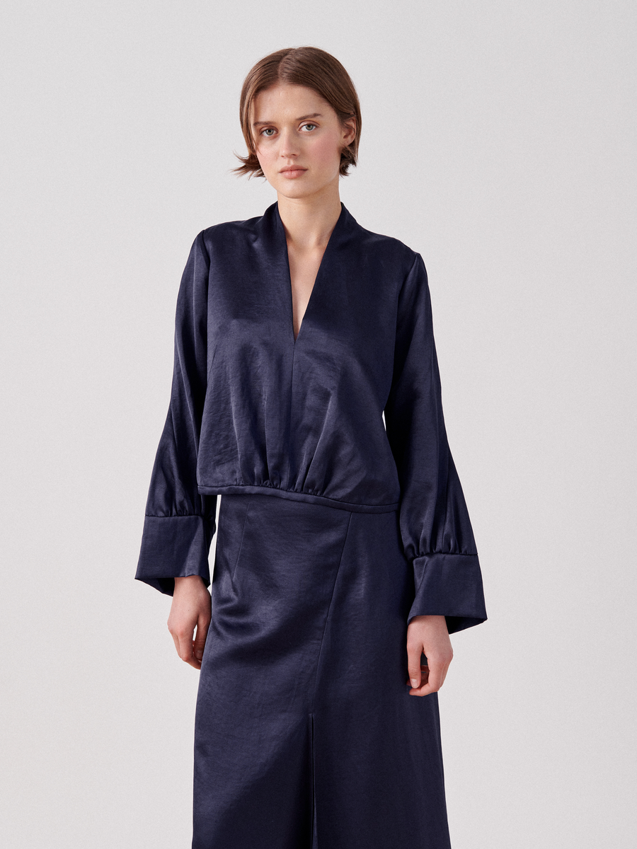 A woman with short hair is wearing a long-sleeved, dark blue linen blend Simple Sacha Top by Zero + Maria Cornejo and a matching skirt, both featuring a v-neckline. She stands against a plain white background with a neutral expression, her hands relaxed by her sides.
