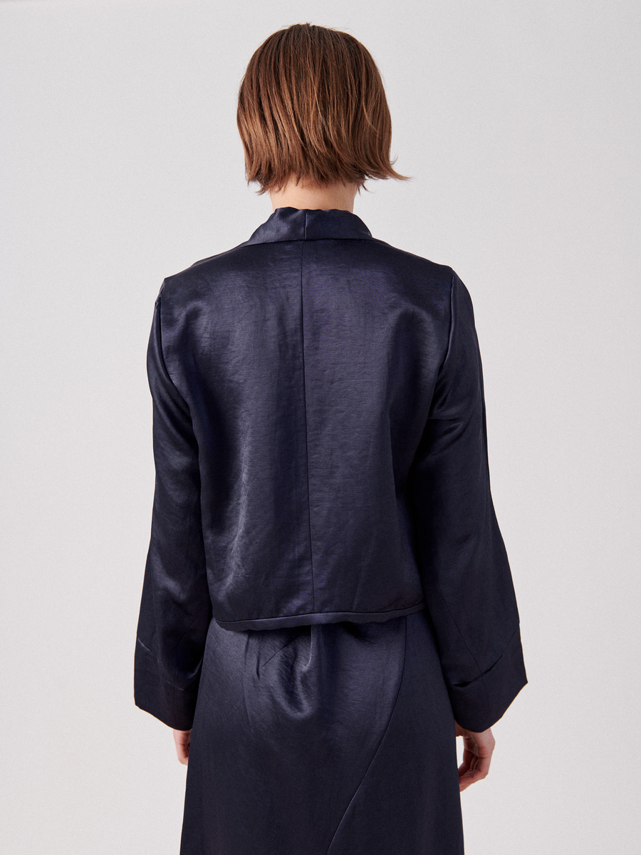 A person with short, light brown hair wearing a dark, silky jacket and matching Simple Sacha Top by Zero + Maria Cornejo stands facing away from the camera against a plain white background. The loose-fitting outfit features long sleeves and a subtle v-neckline.