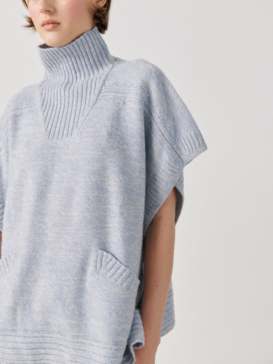 A person wearing a Square Poncho from Zero + Maria Cornejo made from recycled cashmere yarn with a turtleneck and front pockets. The poncho features a textured, ribbed design around the neck and pockets. The background is plain and white.