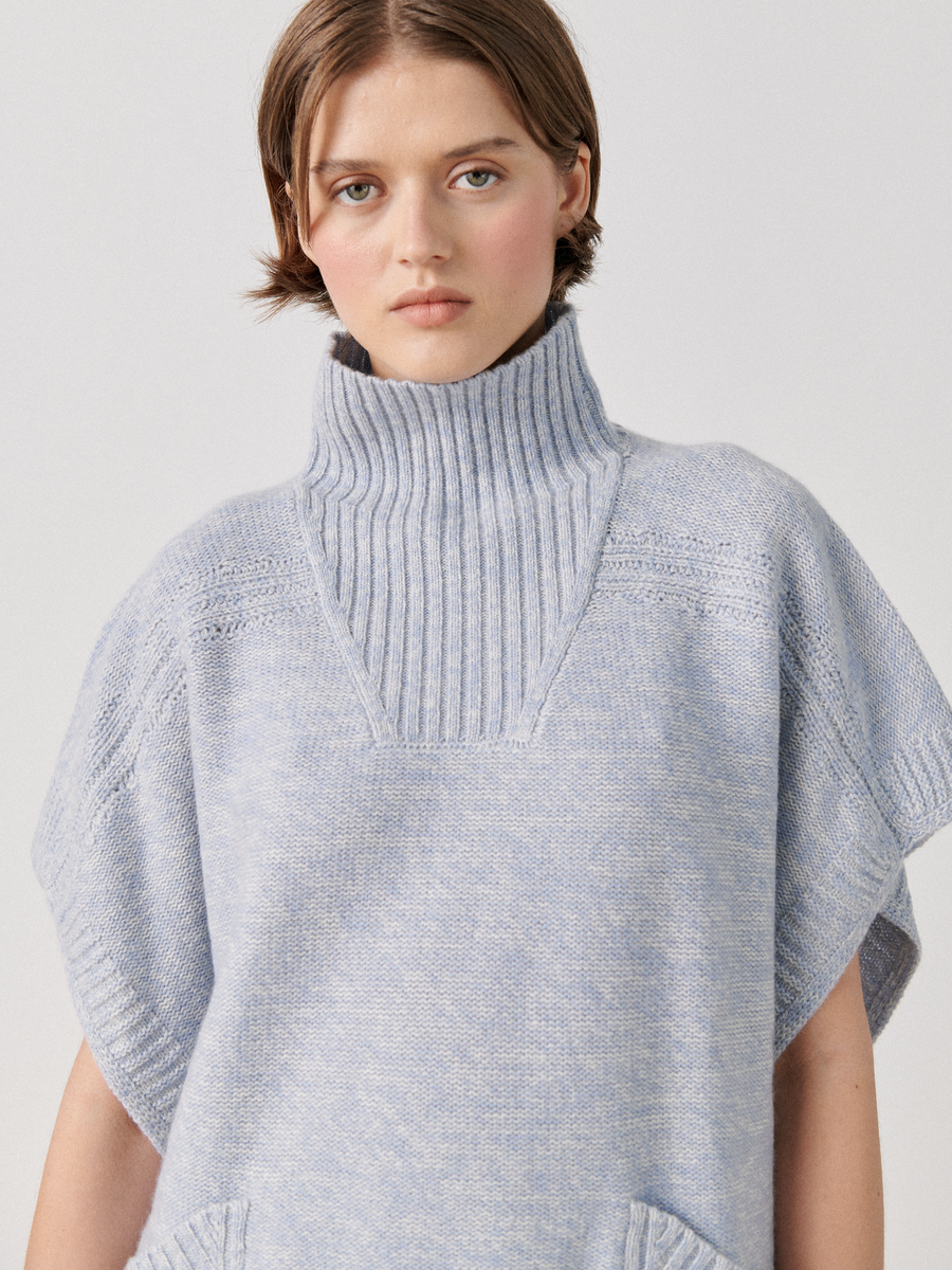 A person stands against a plain background wearing a Square Poncho from Zero + Maria Cornejo. Crafted from recycled cashmere yarn, the poncho is both stylish and eco-friendly. They have short, brown hair and a neutral expression.