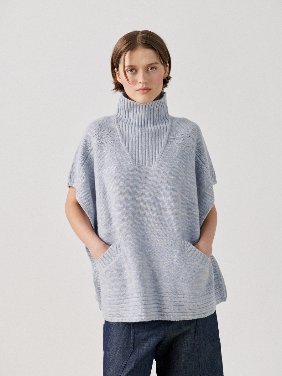 A person with short brown hair stands against a plain background. They are wearing the Zero + Maria Cornejo Square Poncho, a light blue, short-sleeved, high-neck poncho crafted from sustainably-sourced cashmere, featuring two front pockets and dark blue jeans. Their expression is neutral.