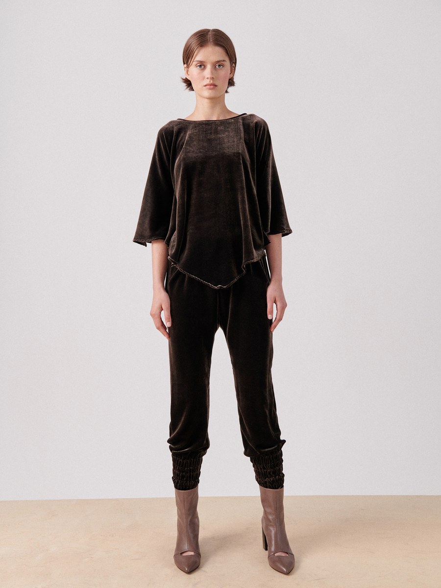 A person stands against a plain background wearing a dark brown, loose-fitting velvet top with elbow-length sleeves and matching luxurious Tri Tabi Pant by Zero + Maria Cornejo featuring a banded elastic waist and hidden pockets. They are also wearing brown ankle boots. The attire has a relaxed, comfortable style.