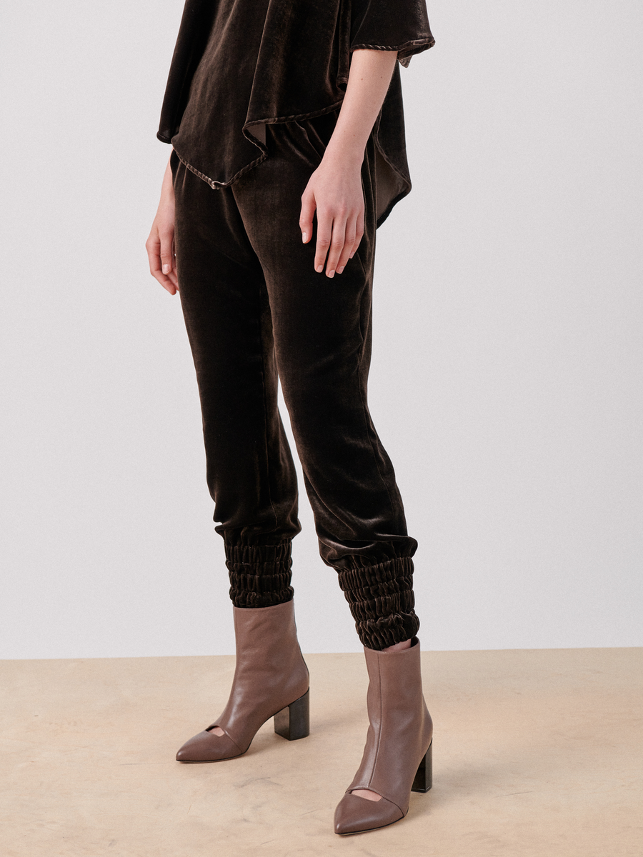 A person is standing against a plain white background, wearing Zero + Maria Cornejo Tri Tabi Pants with elastic cuffs at the ankles and a banded elastic waist, paired with a matching top. They are also wearing stylish brown ankle boots with block heels. The luxurious velvet trousers feature hidden pockets. The person's face is not visible.