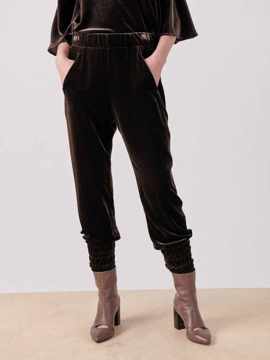 A person is wearing luxurious Zero + Maria Cornejo Tri Tabi Pant with banded elastic ankle cuffs, and their hands are in the hidden pockets. They are also sporting taupe-colored heeled ankle boots. The background is plain and light-colored, and the photo is cropped to show only the lower part of the body.