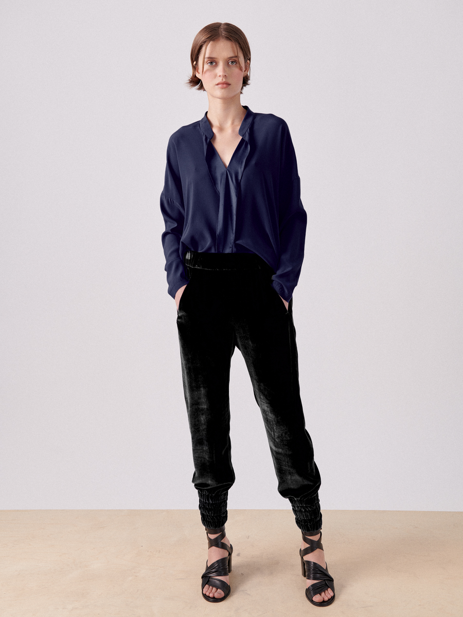 A person with short hair stands against a plain background, wearing a dark blue, long-sleeved blouse and luxurious Tri Tabi Pant by Zero + Maria Cornejo with a banded elastic waist and gathered ankles. Black strappy sandals complete the look as they keep their hands in hidden pockets and maintain a neutral expression.