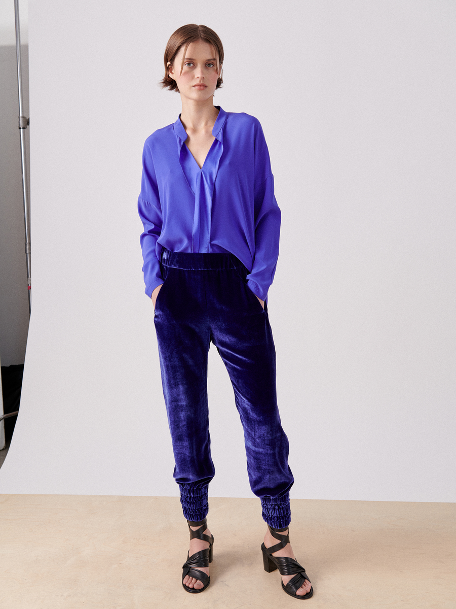 A person with short hair stands against a plain backdrop, wearing a bright blue blouse tucked into luxurious Tri Tabi Pant by Zero + Maria Cornejo with a banded elastic waist. They have their hands in hidden pockets and are wearing black open-toe heels.
