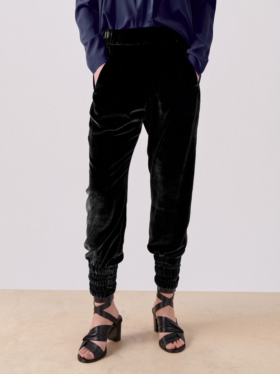 A person is shown from the shoulders down, wearing a dark blue, long-sleeved shirt with hands in the hidden pockets of luxurious velvet Tri Tabi Pant by Zero + Maria Cornejo. The pants have elastic cuffs at the ankles and a banded elastic waist. The person is also wearing black strappy wedge sandals.