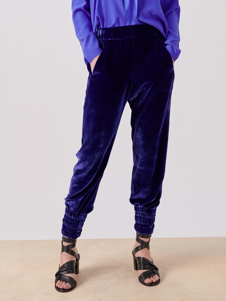 Person wearing a blue long-sleeve blouse, luxurious velvet **Tri Tabi Pant** by **Zero + Maria Cornejo** with gathered cuffs and banded elastic waist, and black strappy high-heeled sandals. The person’s hands are in their hidden pockets, and their face is not visible.