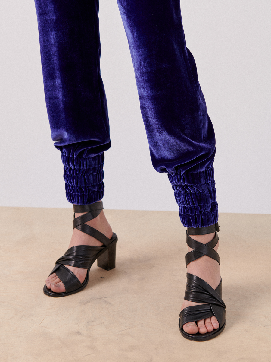 A person is wearing royal blue luxurious velvet Zero + Maria Cornejo Tri Tabi Pant with gathered ankles and black strappy high-heeled sandals. The image captures the lower legs and shoes against a neutral background, showcasing the elegance of the banded elastic waist.