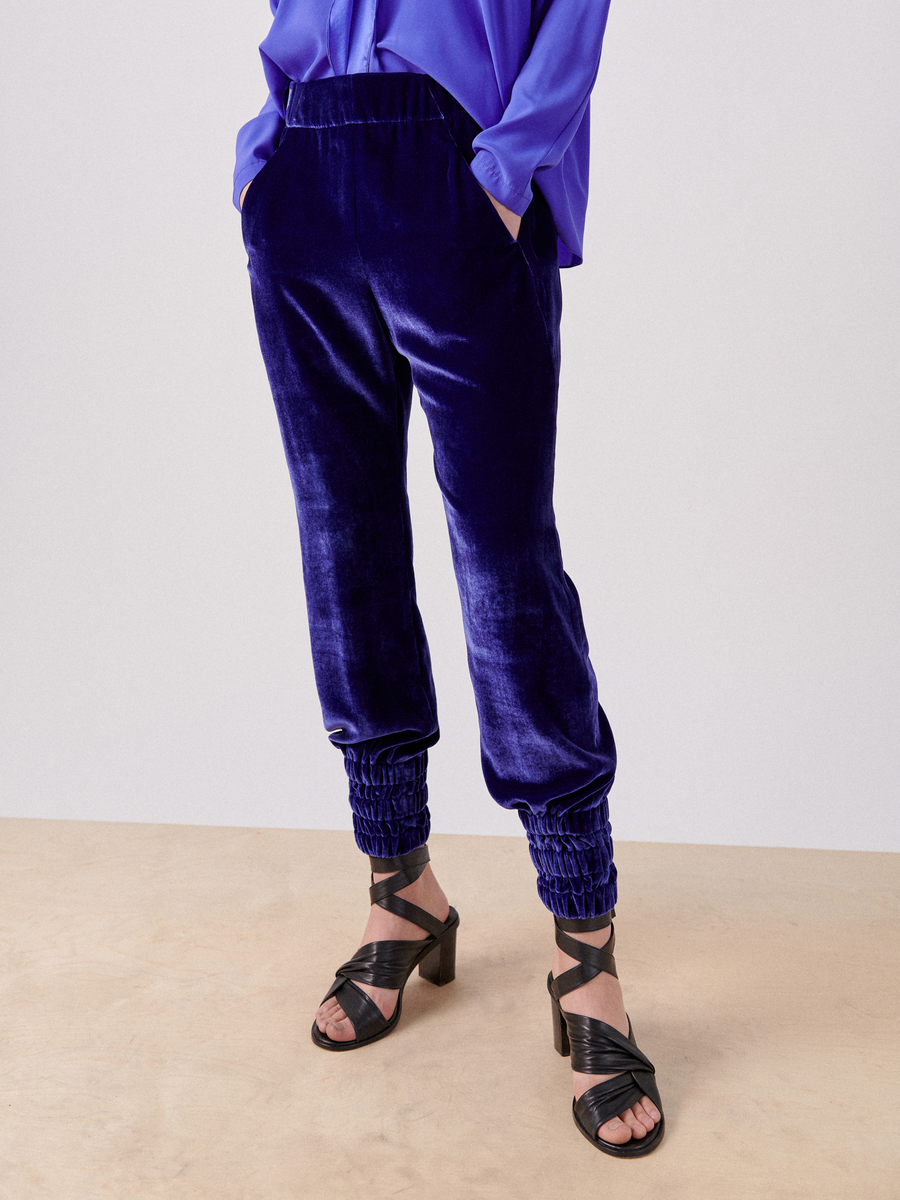 A person stands wearing a bright blue blouse and matching luxurious velvet Tri Tabi Pant by Zero + Maria Cornejo, which feature elasticated ankle cuffs. The pants include a banded elastic waist for added comfort. The individual is also sporting black strappy high-heeled sandals. The background is plain and neutral, with the person's upper body not fully visible.