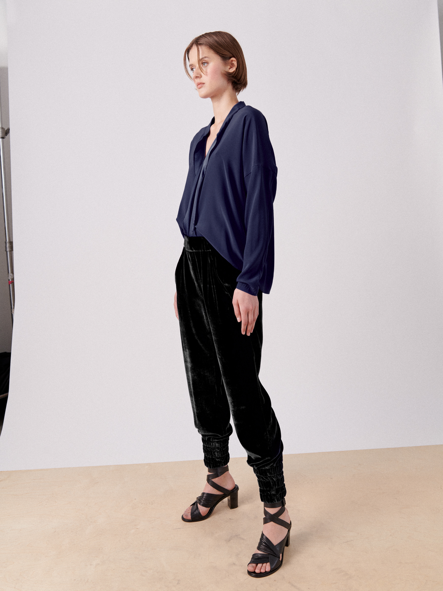 A person with short brown hair is standing against a plain background. They are wearing a loose-fitting navy blue blouse, luxurious velvet Zero + Maria Cornejo Tri Tabi Pant with banded elastic waist, and black strappy heels. The person has a relaxed pose with one hand in their hidden pocket and gazes off to the side.